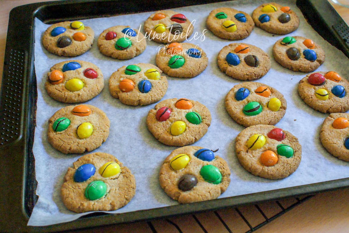 Plaque de cuisson garnie de cookies aux M&M’s tout juste sortis du four, disposés sur du papier sulfurisé, aux couleurs vives et légèrement craquelés – ambiance gourmande de biscuits faits maison